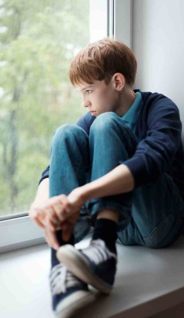 young teenage boy sitting at the window looking sad