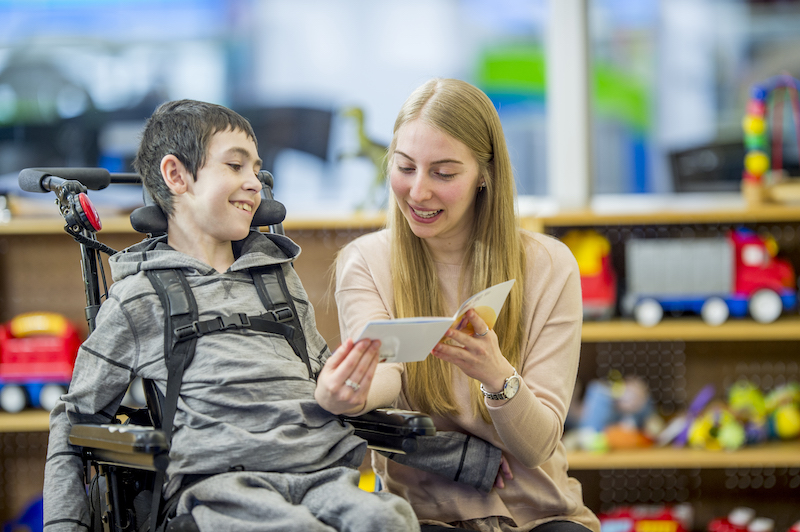 A boy in a wheelchair is indoors in his classroom. He is being assisted by his classroom helper. She is reading a story to him.