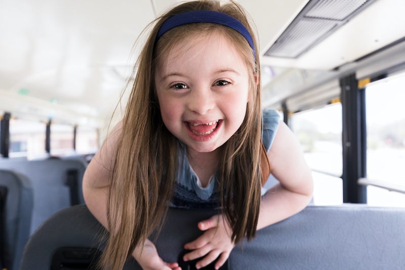 A young girl with Down Syndrome leans over bus seat to smile at camera.  She is enjoying herself.