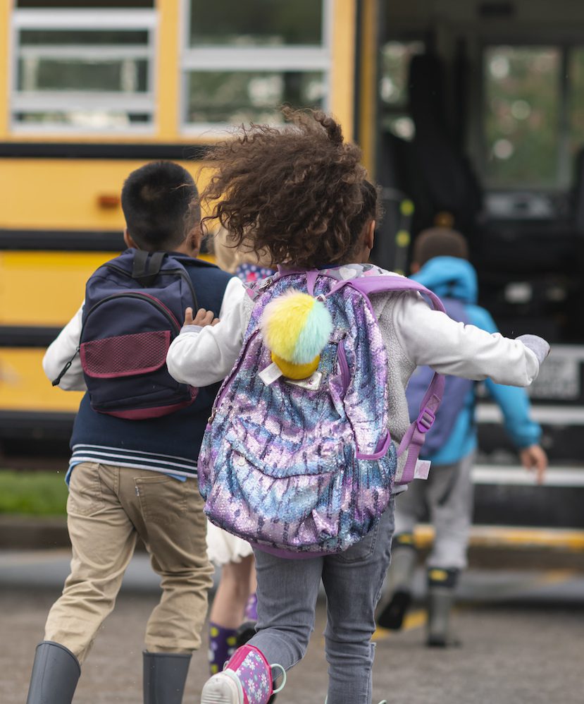 Diverse group of happy children getting on school bus A multi-ethnic group of elementary age children are getting on a school bus. The kids' backs are to the camera. They are running towards the school bus which is parked with its door open. It's a rainy day and the kids are wearing jackets, rain boots and backpacks.
